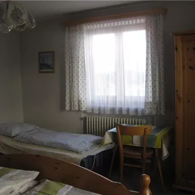 Ein einfaches Zimmer mit zwei Betten, einem Tisch und einem Stuhl. Das Fenster lässt viel Licht herein und die Wände sind hell gestrichen.
