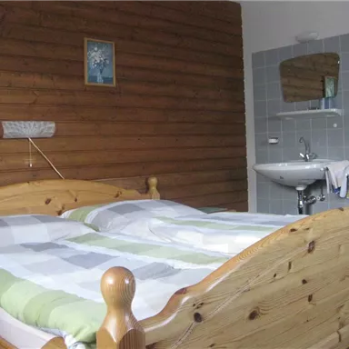 Ein einfaches Schlafzimmer mit einem Holzbetten und einer hellen Decke. Im Hintergrund befindet sich ein Waschbecken und ein Spiegel.