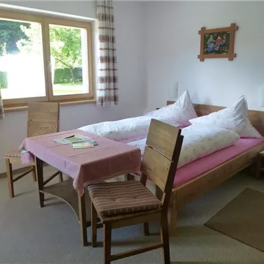 Ein gemütliches Zimmer mit einem Doppelbett und einem Tisch. Die Fenster lassen viel Licht herein und der Raum wirkt einladend und freundlich.