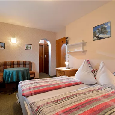 Ein gemütliches Zimmer mit zwei Betten, einer Sitzecke und bunten Kissen. Die Wände sind mit Bildern geschmückt, und es gibt eine warme, einladende Atmosphäre.