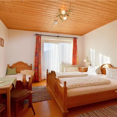 A bright bedroom with two single beds and a small table. Large windows let in plenty of light, and the furniture is made of light wood.