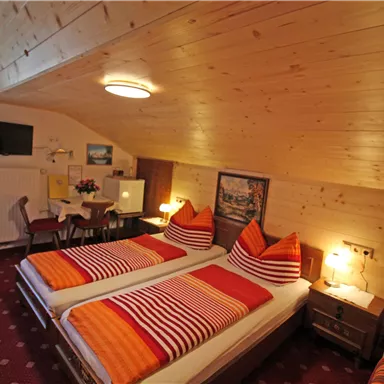 Ein gemütliches Schlafzimmer im Dachgeschoss mit zwei Einzelbetten und bunten Kissen. Der Raum ist mit Holzwänden, einem Fernseher und einem kleinen Tisch ausgestattet.