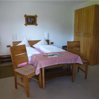 Ein einfaches Schlafzimmer mit einem Bett, einem Tisch und Stühlen. Der Raum hat einen neutralen Farbton und ein Fenster mit Vorhängen.