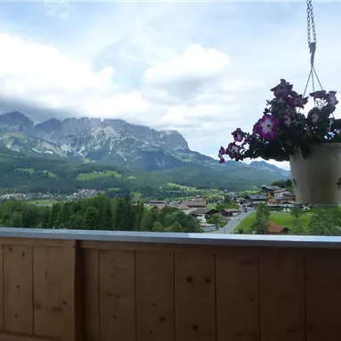 Eine schöne Landschaft mit Bergen im Hintergrund und einer hängenden Pflanzenampel. Im Vordergrund ist ein Balkon aus Holz sichtbar.