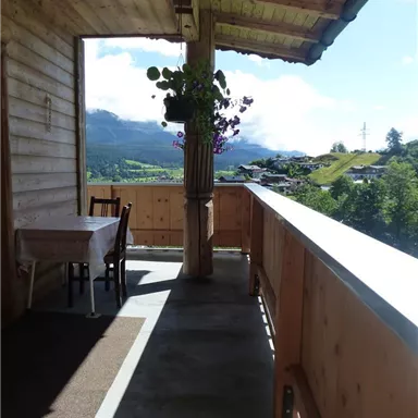Ein Balkon mit Holzgeländer und einer Tisch-Stuhl-Kombination. Im Hintergrund sind Hügel und Berge sichtbar, unter einem klaren Himmel.