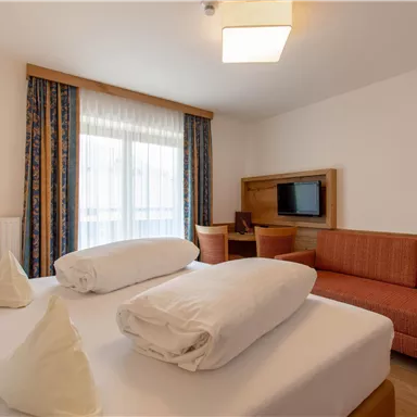 Ein modernes Zimmer mit einem Doppelbett, zwei Kissen und einem gemütlichen Sofa. Das Zimmer hat große Fenster, die viel Tageslicht hereinlassen.