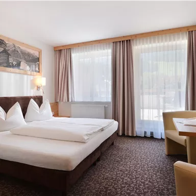 A modern hotel room with a double bed and two large windows. The room is cozy and equipped with a table and a television.