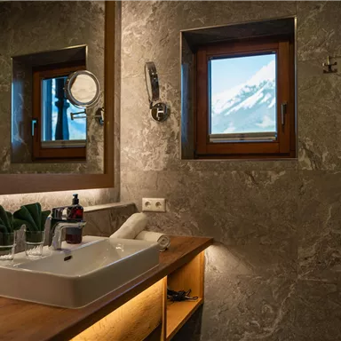 Ein modernes Badezimmer mit einem eleganten Waschbecken und Holzregalen. Durch das Fenster ist eine beeindruckende Berglandschaft zu sehen.