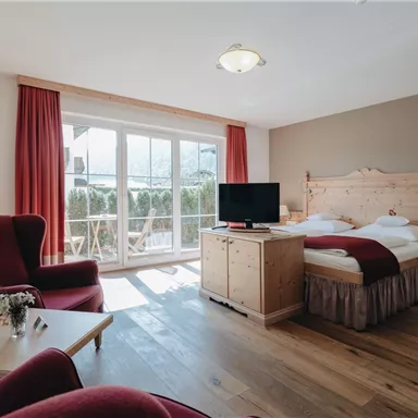 Ein gemütliches Zimmer mit einem Doppelbett und einem Fernseher. Große Fenster lassen viel Licht herein und bieten einen Blick auf den Balkon.