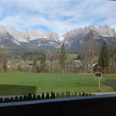 An impressive mountain landscape with snow-covered peaks and a green valley. In the foreground, trees and meadows rise.