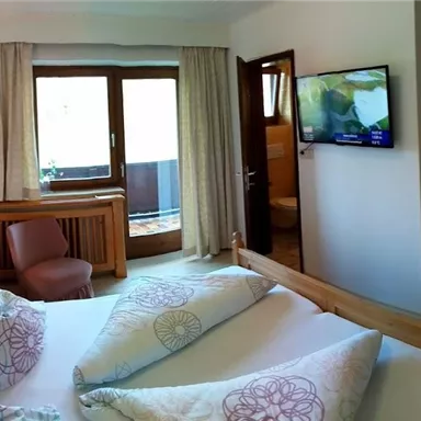 A cozy hotel room with a large window and an outside view. There is a double bed, a television, and wooden furniture for a warm atmosphere.