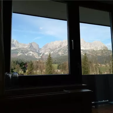 A beautiful view of majestic mountains through a window. There is a clear blue sky and green forests in the foreground.