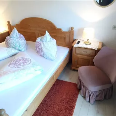 A cozy bedroom with a wooden bed and two pillows. Next to the bed is a chair and a small bedside lamp.