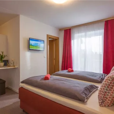 Ein gemütliches Schlafzimmer mit zwei Betten, einem Fernseher und einem kleinen Tisch. Große Fenster lassen viel Licht herein, und rote Vorhänge verleihen dem Raum Wärme.