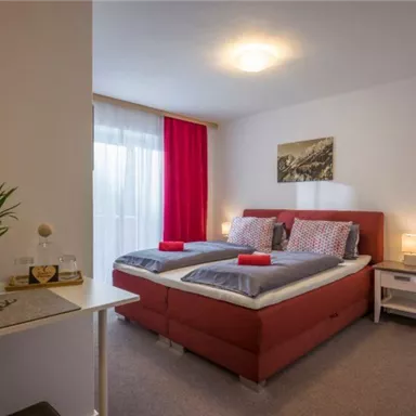 Ein gemütliches Schlafzimmer mit zwei Betten, roten Akzenten und einer großen Fensterfront. Der Raum ist gut beleuchtet und modern eingerichtet.