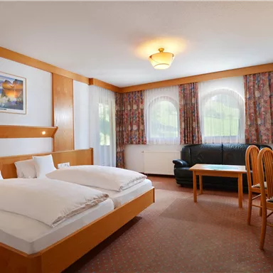 Ein gemütliches Hotelzimmer mit zwei Betten, einem Fernseher und einer Sitzecke. Große Fenster sorgen für viel Tageslicht.