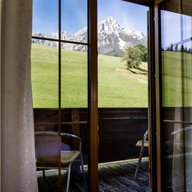 Ein Blick aus einem Zimmerfenster auf grüne Wiesen und die schneebedeckten Berge im Hintergrund. Die Umgebung wirkt ruhig und einladend.