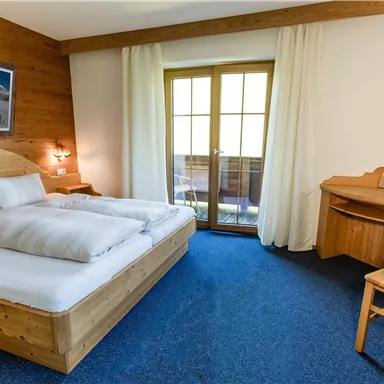 Ein gemütliches Hotelzimmer mit einem Doppelbett und einem Holzschreibtisch. Große Fenster bieten Blick auf einen Balkon mit natürlichem Licht.