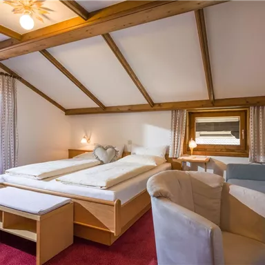 Ein gemütliches Schlafzimmer mit einem Doppelbett und einer Sitzecke. Der Raum hat helle Wände und ein großes Fenster, das Tageslicht hereinlässt.