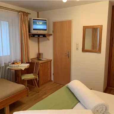 Ein gemütliches Hotelzimmer mit einem Bett, Holzregalen und einem Fernseher. Im Zimmer befinden sich ein Tisch mit Stühlen und ein Fenster mit Vorhängen.