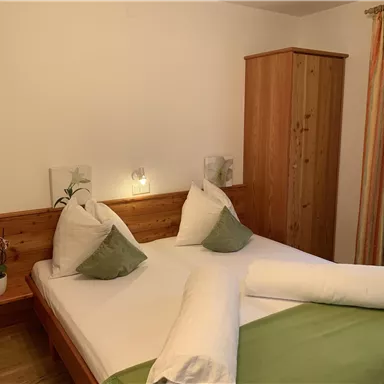 Ein gemütliches Schlafzimmer mit zwei Betten, Kissen und einer Holzkommode. Das Zimmer hat helle Wände und Dekorationen in sanften Farben.