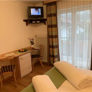 Ein gemütliches Hotelzimmer mit einem Tisch, Stühlen und einem Fernseher an der Wand. Große Fenster sorgen für viel Tageslicht.