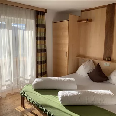 Ein gemütliches Schlafzimmer mit einem großen Bett und hellen, natürlichen Holzfarben. Vor einem Fenster hängen leichte Vorhänge, und eine grüne Decke liegt auf dem Bett.