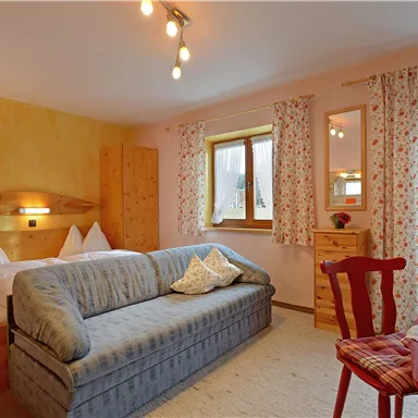 Ein gemütliches Zimmer mit einem Sofa, einem Tisch und zwei Betten. Die Wände sind in sanften Farben gestrichen, und große Fenster lassen viel Licht herein.