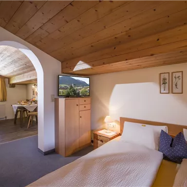 A cozy bedroom with a wooden ceiling and two beds. In the background, you can see a living area with a dining table and a TV.
