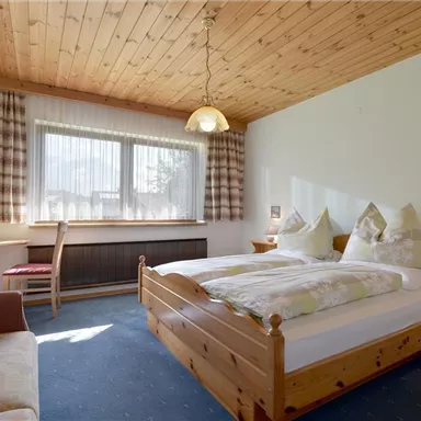 A cozy bedroom with a wooden ceiling and a large window. The room is furnished with a double bed, a sofa, and a table.