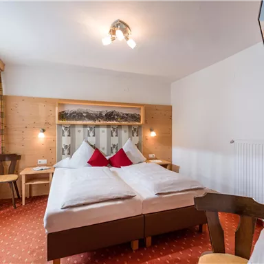 A cozy bedroom with a double bed, decorated with red cushions. The walls are made of wood, and the room has plenty of natural light through a large window.
