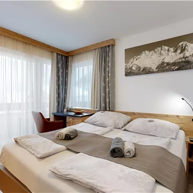 A bright hotel room with a double bed and wooden furniture. Large windows offer a beautiful view of the mountains.