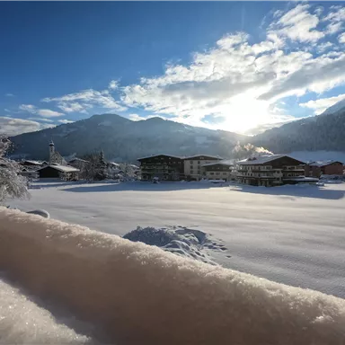 Eine winterliche Landschaft mit schneebedeckten Feldern und Bergen. Die Sonne scheint durch die Wolken und erleuchtet die Szene.