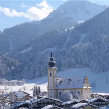 Eine malerische Winterlandschaft mit schneebedeckten Bergen. Im Vordergrund steht eine schöne Kirche umgeben von traditioneller Architektur.
