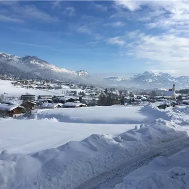 Eine malerische Winterlandschaft mit schneebedeckten Hügeln und kleinen Häusern. Der Himmel ist klar und die Berge sind in der Ferne sichtbar.