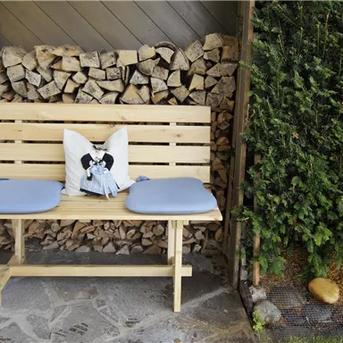 Eine einfache Holzbank mit grauen Kissen und einem Kissen mit einem Muster sitzt vor einem Stapel Holz. Im Hintergrund sind grüne Pflanzen zu sehen.