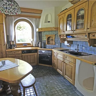 Eine gemütliche Küche im Landhausstil mit Holzschränken und blauen Fliesen. Ein runder Tisch und ein Fenster lassen viel Licht herein.