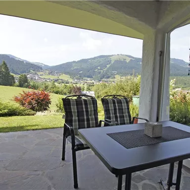 Eine gemütliche Terrasse mit einem Tisch und zwei Stühlen. Im Hintergrund sind grüne Hügel und eine wunderschöne Landschaft zu sehen.