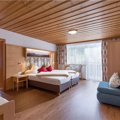 A modern bedroom with wooden paneling and a large window. There is a double bed with red pillows and a cozy sofa.