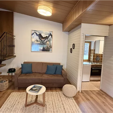 A cozy living room with a sofa, two cushions, and a small table. On the wall hangs a picture of a mountain landscape.