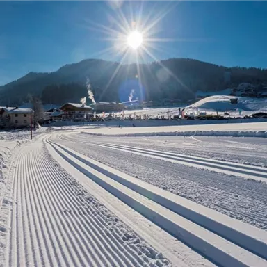 A snowy winter landscape with clear ski trails in the foreground. The sun shines over the mountains and the village in the background.