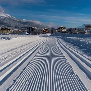 A snow-covered street with clear tracks in the snow and a small snowman on the left side. In the background, snowy mountains and a church can be seen.