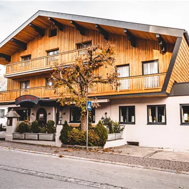 A traditional wooden house with a balcony and large windows. In front of the building, there are trees and there is a quiet street.