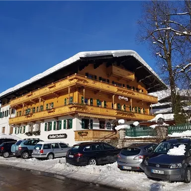 Ein charmantes, schneebedecktes Haus im alpinen Stil mit holzverzierten Balkonen. Umgeben von einer winterlichen Landschaft und geparkten Autos.
