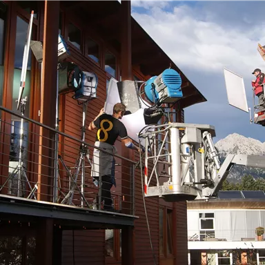 Eine Person arbeitet an einer Film- oder Fotoausrüstung auf einem Balkon. Im Hintergrund sind Berge und eine Wolkenlandschaft zu sehen.