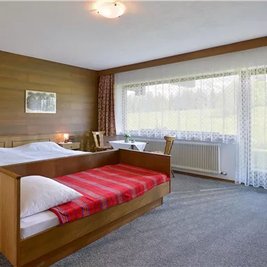 Ein einfaches Schlafzimmer mit zwei Betten und einem Holzmobiliar. Große Fenster bieten viel Tageslicht und Blick auf die Umgebung.