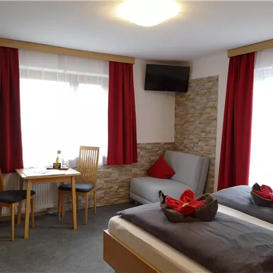 Ein gemütliches Zimmer mit zwei Betten, einem Sofa und einem Tisch. Die Fenster lassen viel Tageslicht herein und die roten Vorhänge verleihen eine warme Atmosphäre.