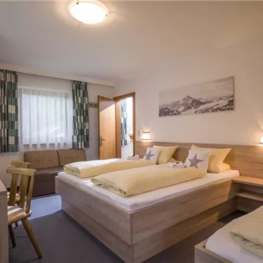 Ein gemütliches Zimmer mit zwei Betten, einem Kleiderschrank und einem Sofa. Helle Farben und natürliche Lichtquellen schaffen eine einladende Atmosphäre.