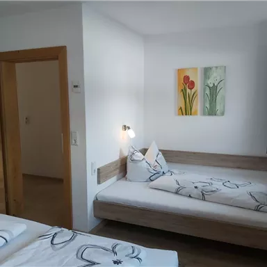 Ein helles Zimmer mit zwei Betten und Blumendekor. An der Wand hängen farbenfrohe Bilder, und ein Fenster lässt viel Licht herein.