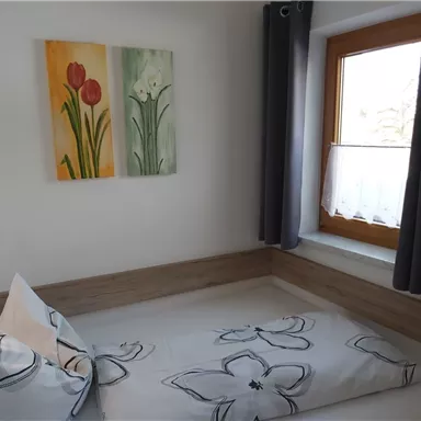 Ein gemütliches Schlafzimmer mit einem einzelnen Bett und floralem Bettzeug. An der Wand hängen bunte Blumenbilder, und ein Fenster lässt viel natürliches Licht herein.
